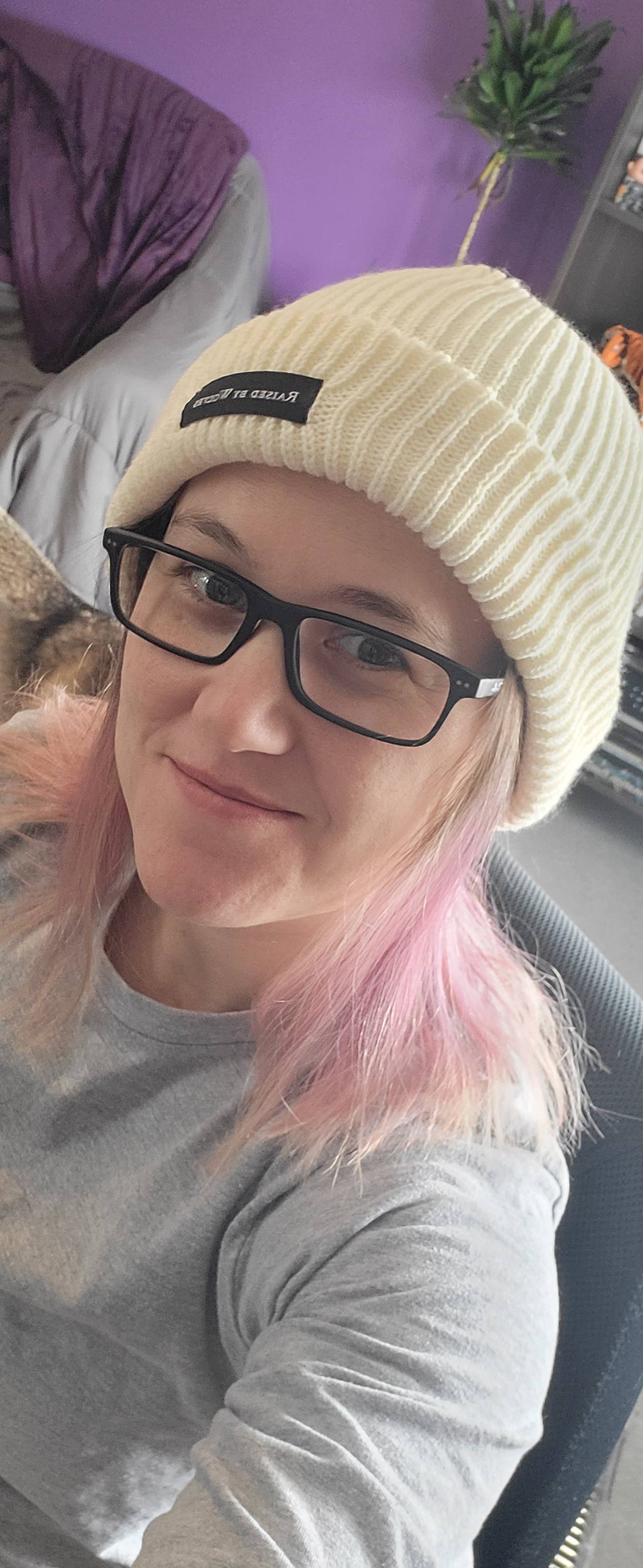 Person wearing a beige beanie and glasses with pink hair, sitting indoors.