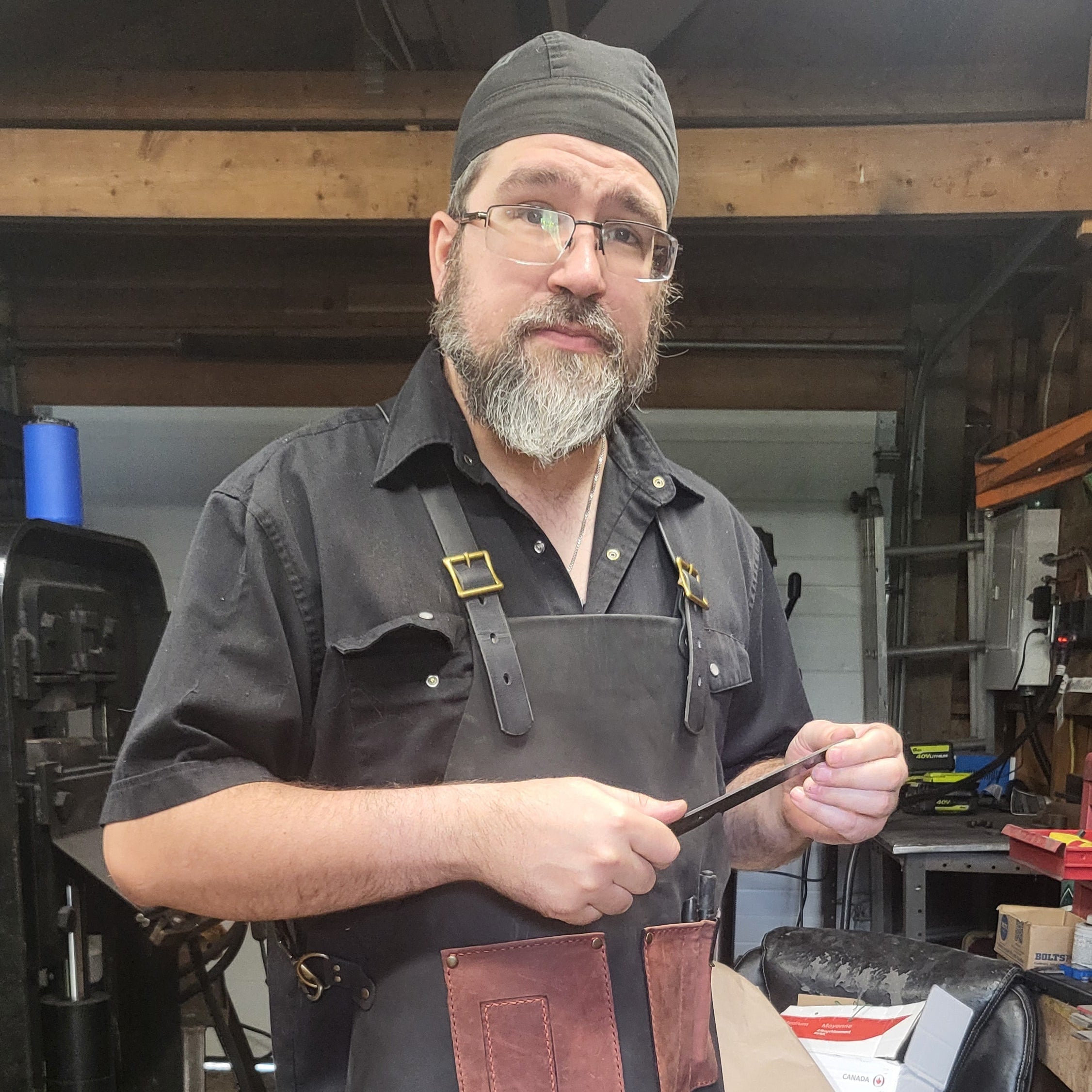 Man in a leather apron holding a knife blank in a workshop setting