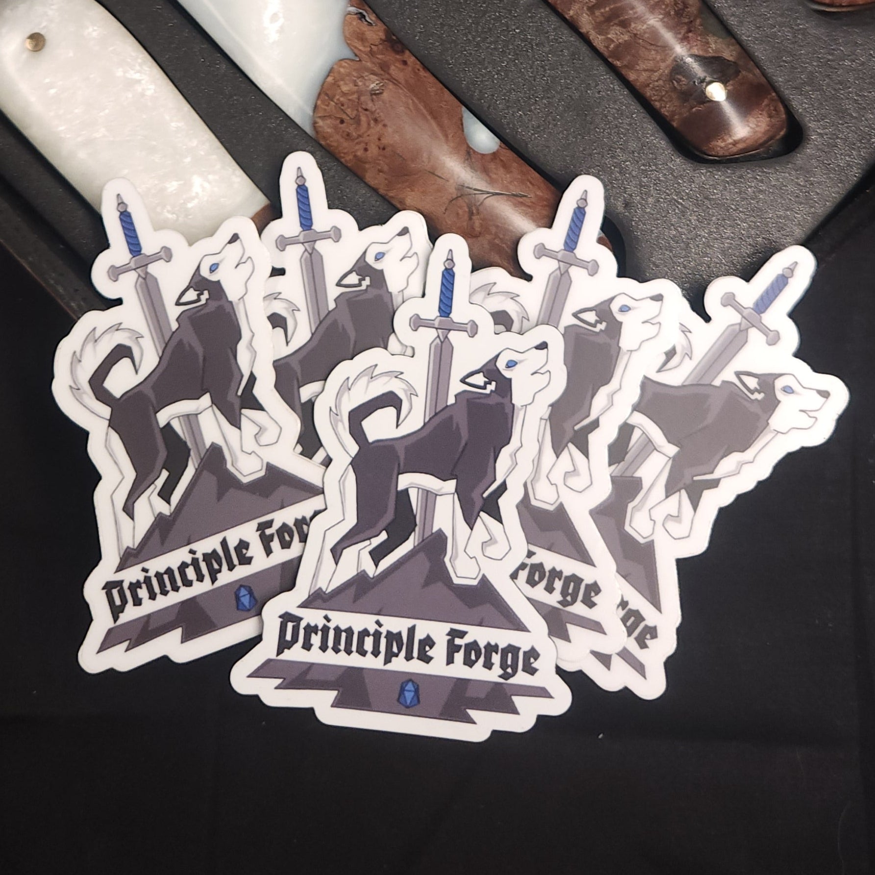 Sticker of the Principle Forge logo featuring a husky on a mountain with a sword and the words 'Principle Forge' sitting on the handles of a set of Damascus steak knives