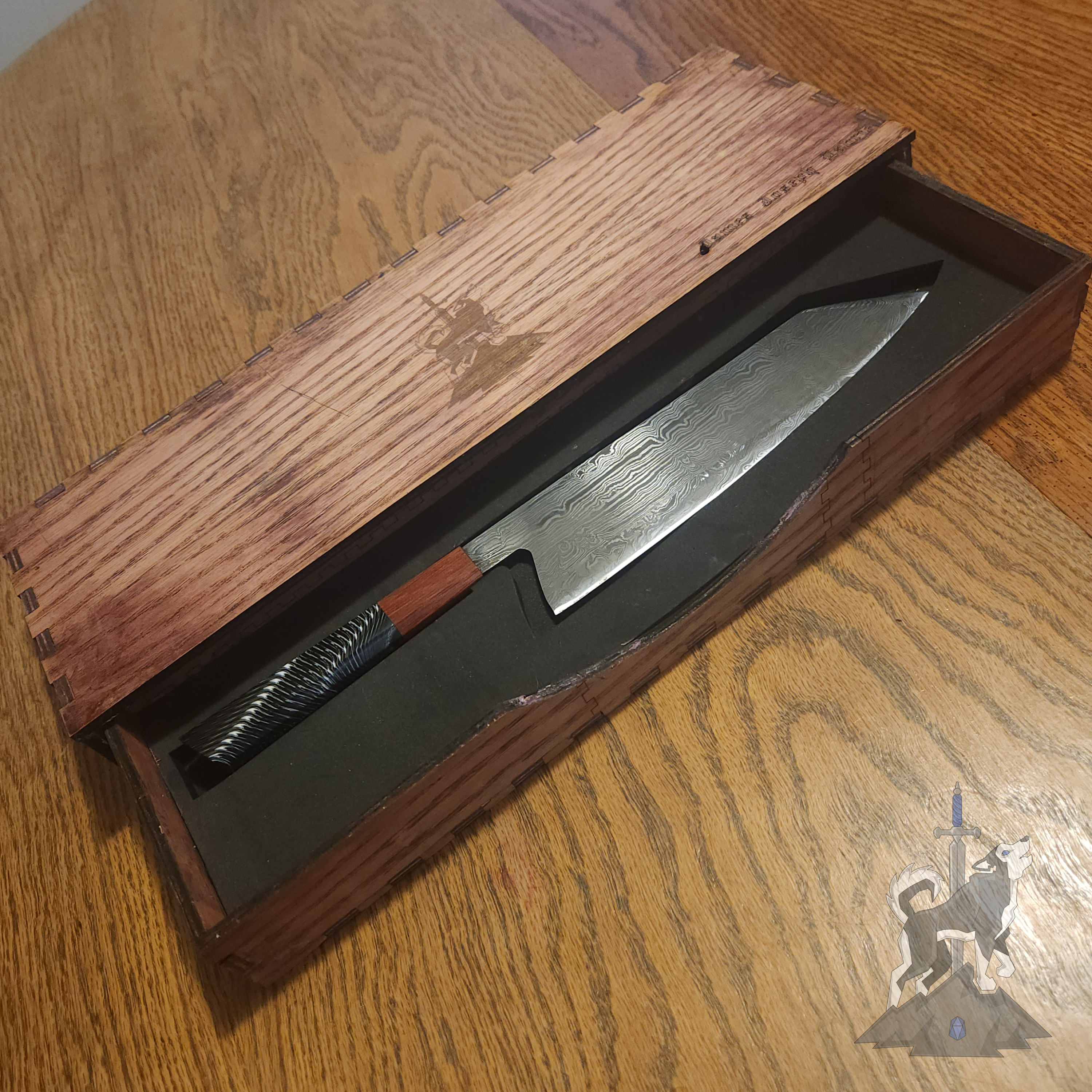 Chef knife with Damascus pattern in its wooden storage box with laser engraved Principle Forge logo on a dark wooden surface.