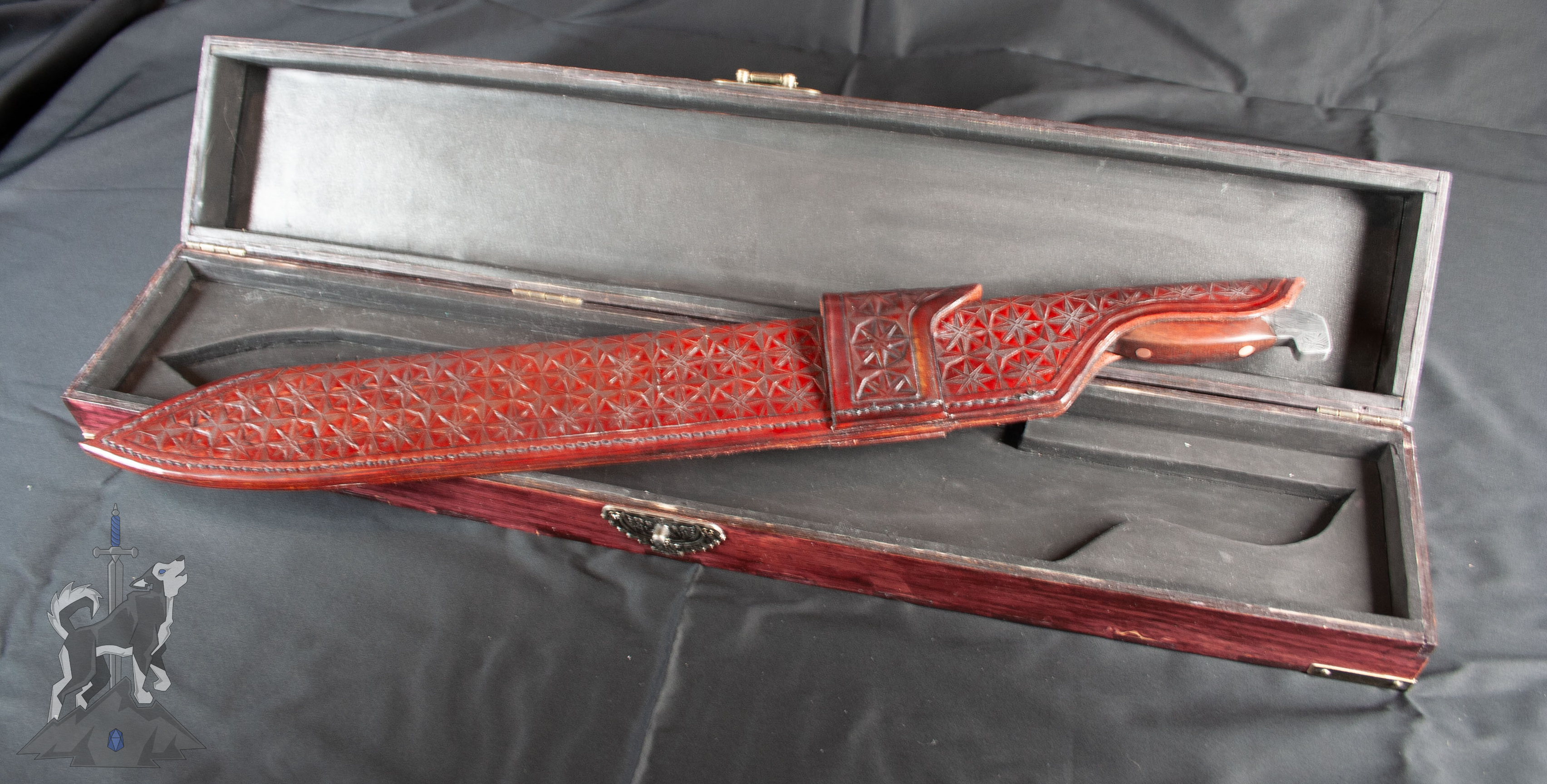 A Damascus kukri hybrid blade in its matching leather sheath with hand tooled star patterns and belt loop sitting on its open wooden storage box on a dark background