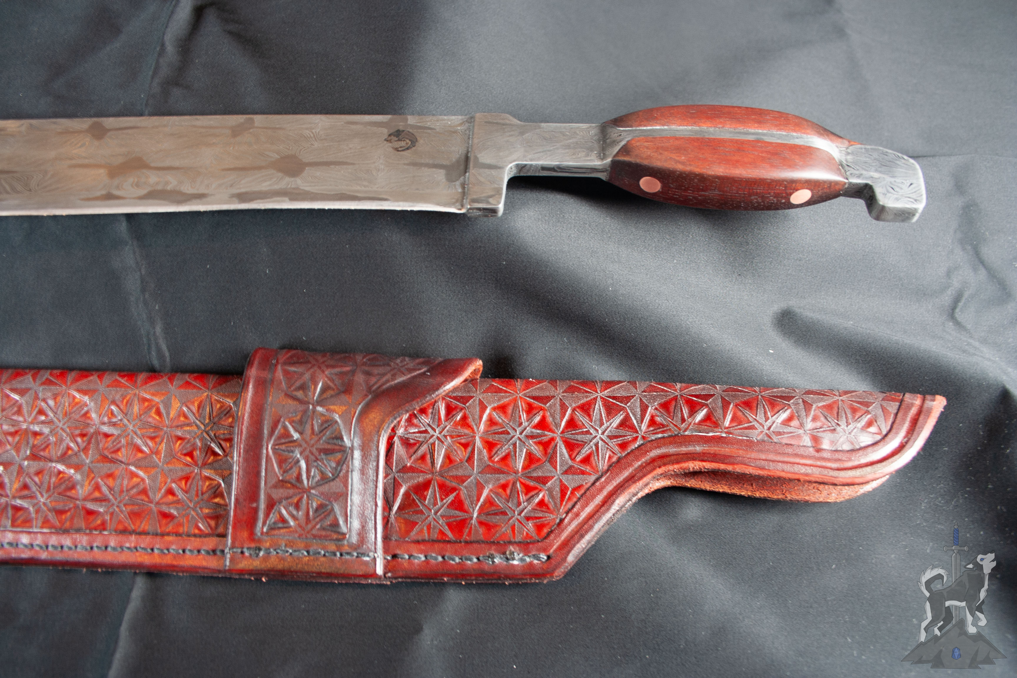 Detail shot of a Damascus kukri hybrid blade showcasing the wooden handle and part of the leather sheath with hand tooled star patterns and belt loop on a dark background