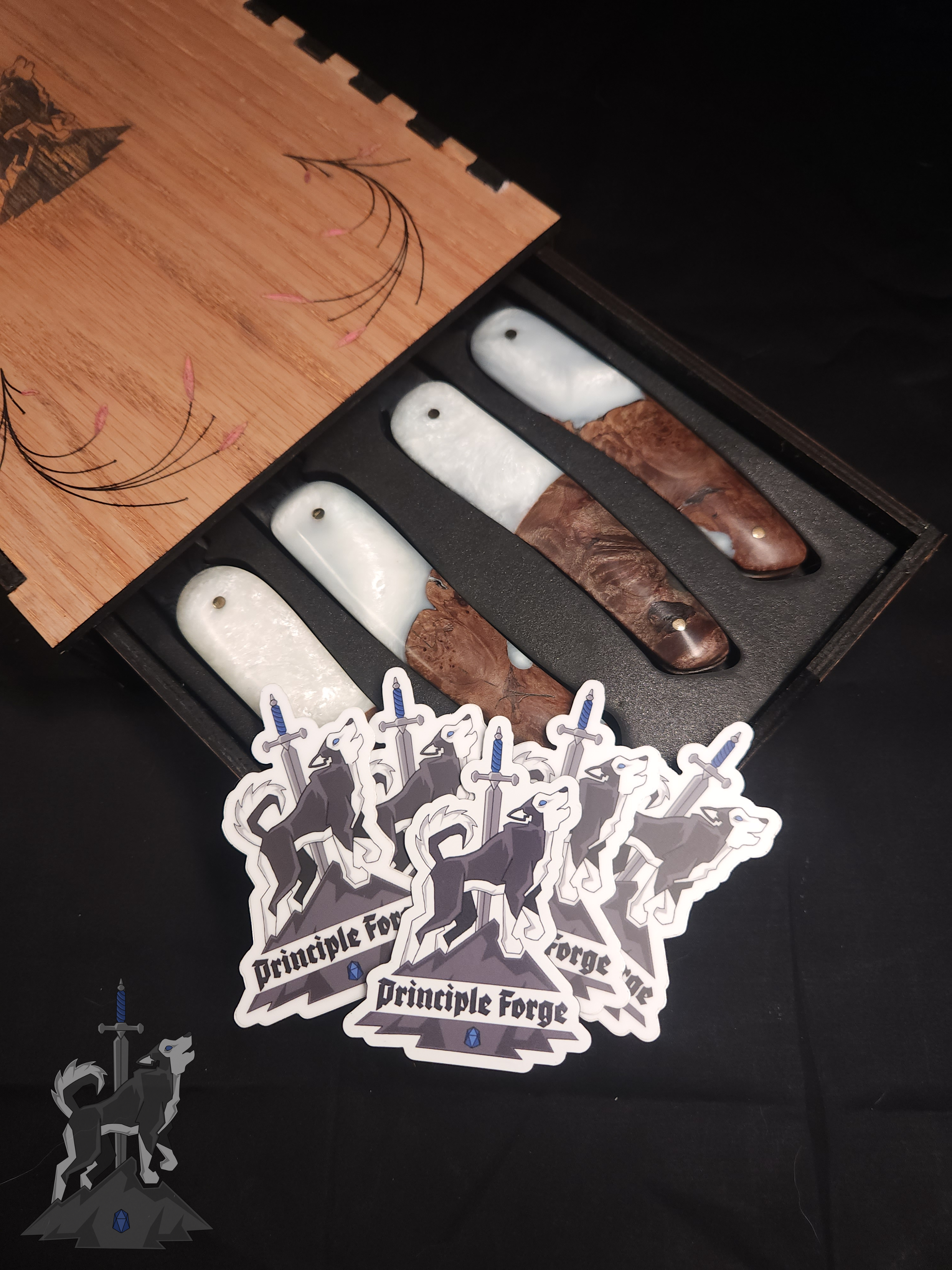 Four feather patterned Damascus steak knives with white and wooden handles seated in their wooden storage box on a dark background with only the ends of the handles visible and several Principle Forge logo stickers sitting on them