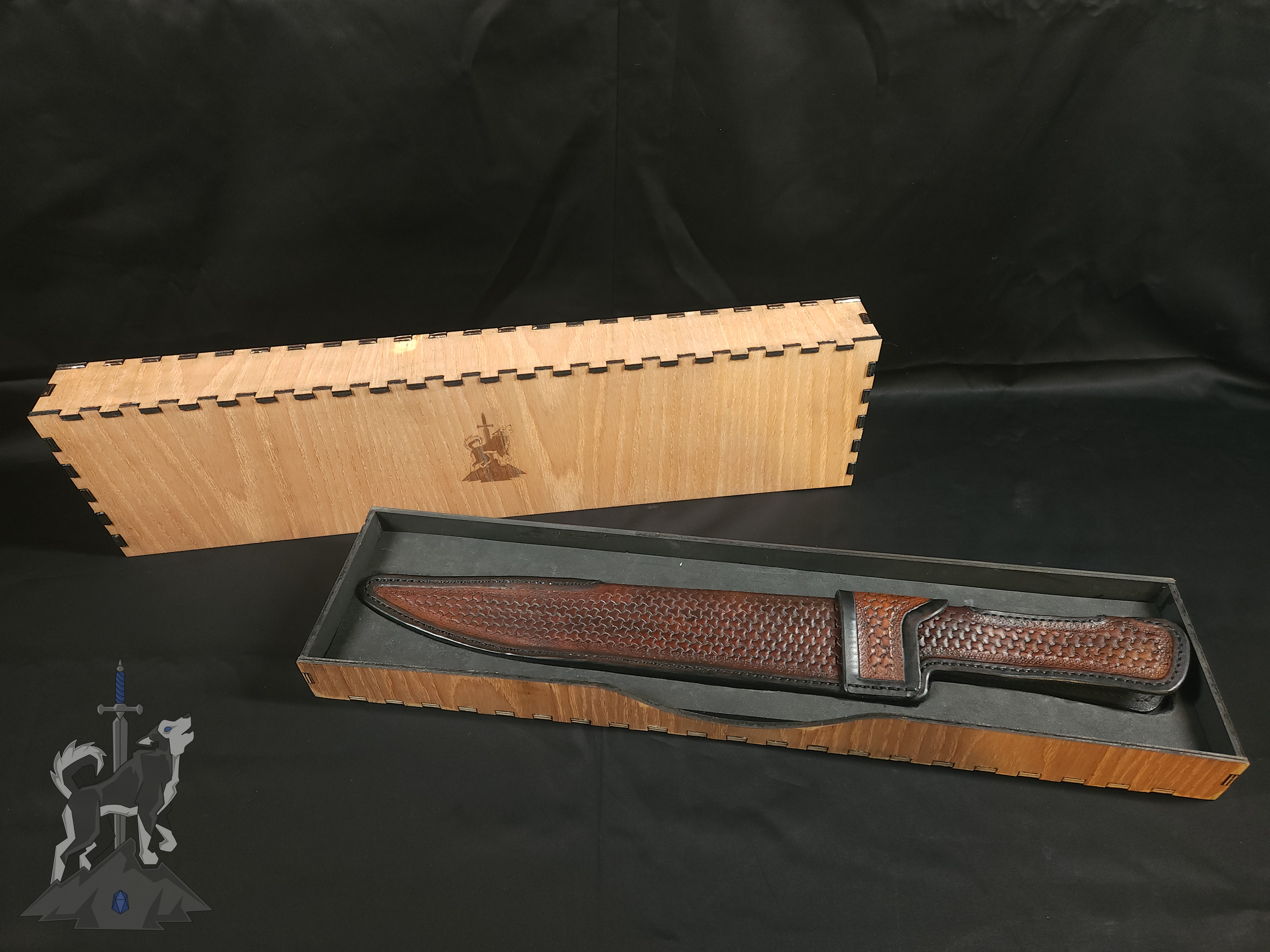 A custom Damascus takedown bowie in its hand tooled leather sheath sitting in its wooden storage box featuring a laser engraved Principle Forge logo.