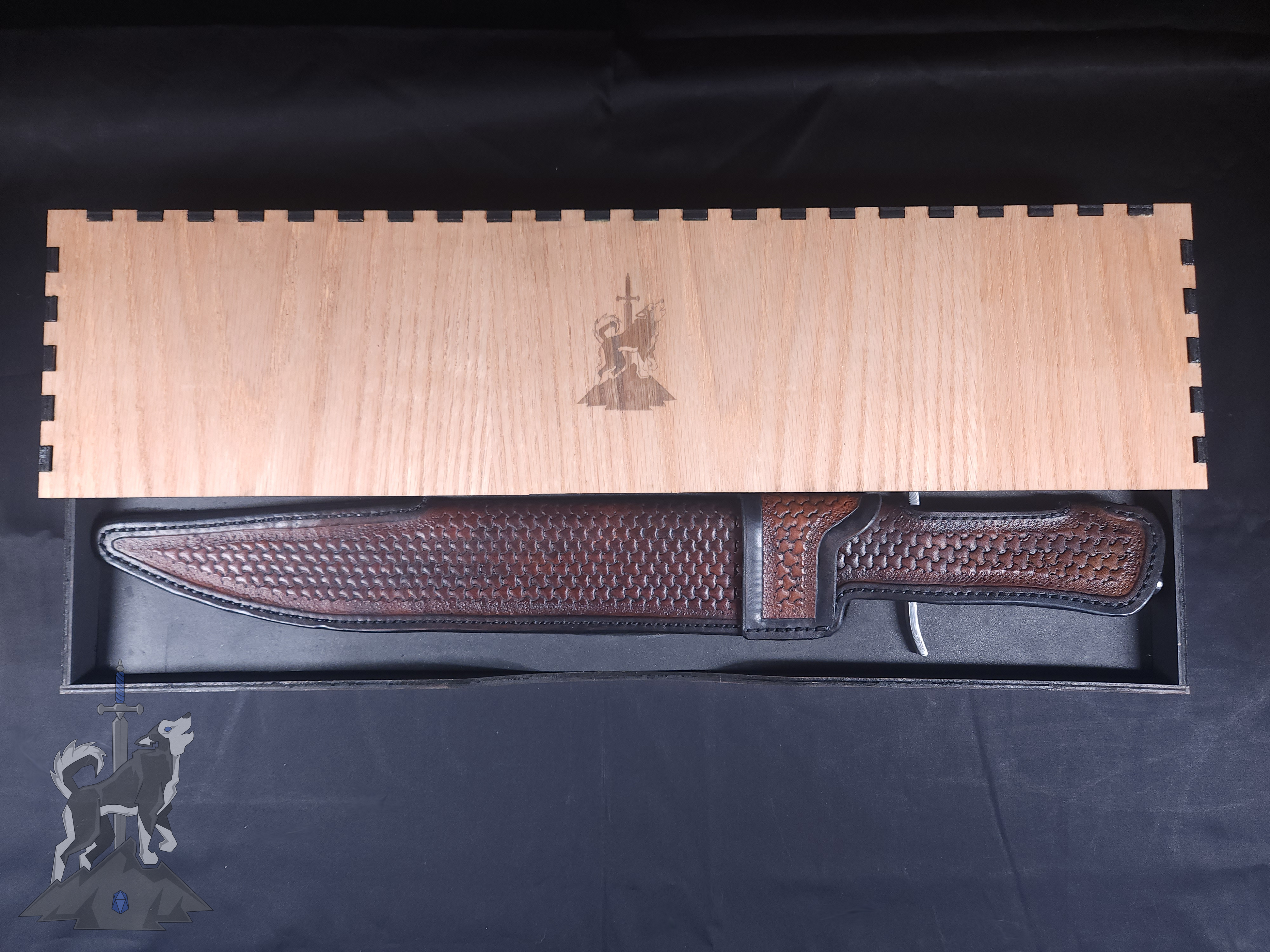 A custom Damascus takedown bowie in its hand tooled leather sheath sitting in its wooden storage box featuring a laser engraved Principle Forge logo.