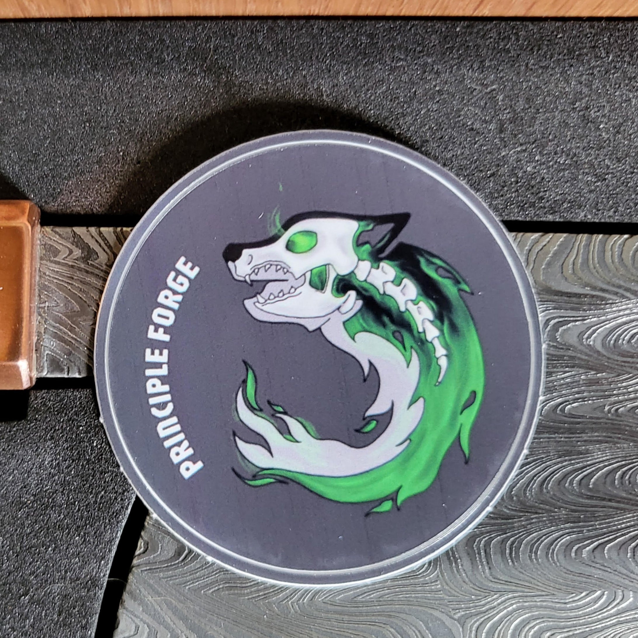 Round sticker featuring a husky skull wrapped in a green and black fireball sitting on a Damascus chef knife